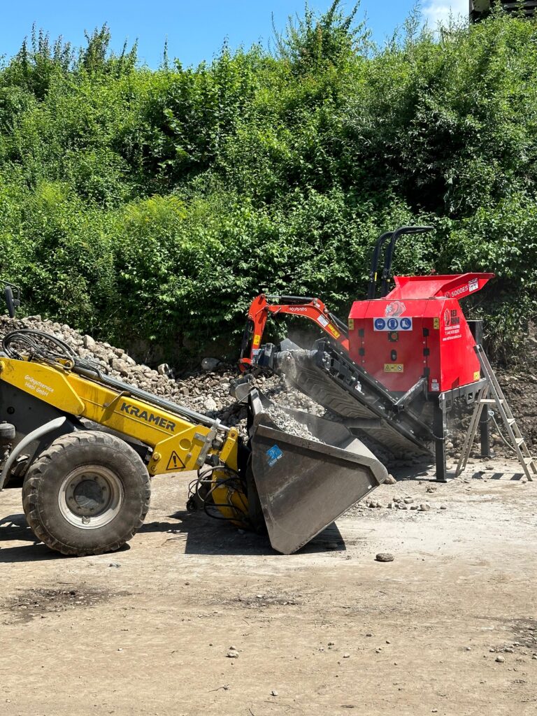 A yellow front-end loader is positioned to the left, using its large bucket to dump broken concrete or rubble into a Red Rhino mobile crushing machine on the right. A large pile of rubble is behind the equipment, with a dense background of green foliage.