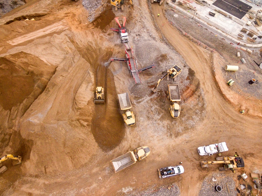 An aerial shot of a large construction or quarry site, showing several dump trucks, excavators, and other heavy machinery moving and sorting large piles of earth and gravel.
