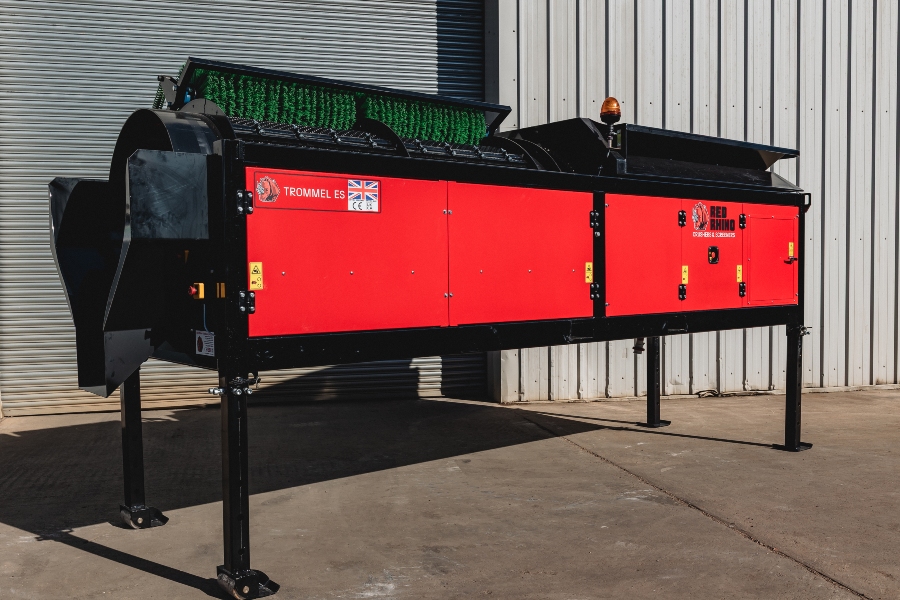 Full outdoor side view of the Red Rhino Electric Trommel screening machine, showing the bright red body panels, the black steel frame, and the four height-adjustable support legs with stabilizer feet on a concrete surface.
