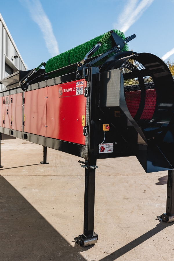 Close-up, angled outdoor shot of the Red Rhino Electric Trommel screening machine, showing the bright red control panel housing, black steel frame, height-adjustable support legs, and the green spring-tensioned cleaning brush on top of the rotating cylindrical screen.