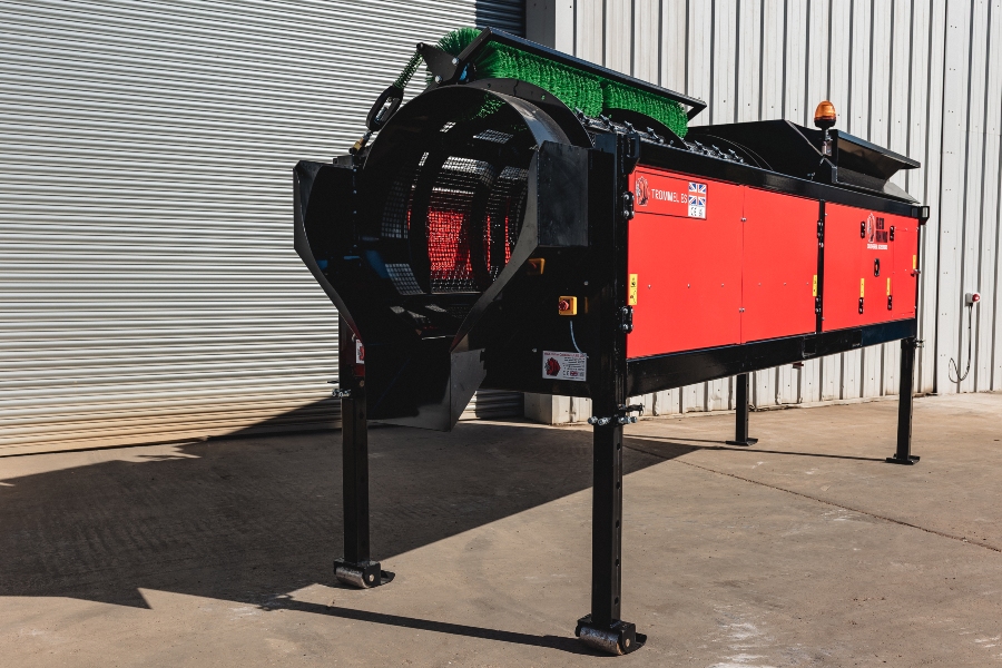 Outdoor full side view of the Red Rhino Electric Trommel screening machine, featuring the bright red body, black steel frame, height-adjustable legs with small wheels, and the green cleaning brush visible above the cylindrical screening drum